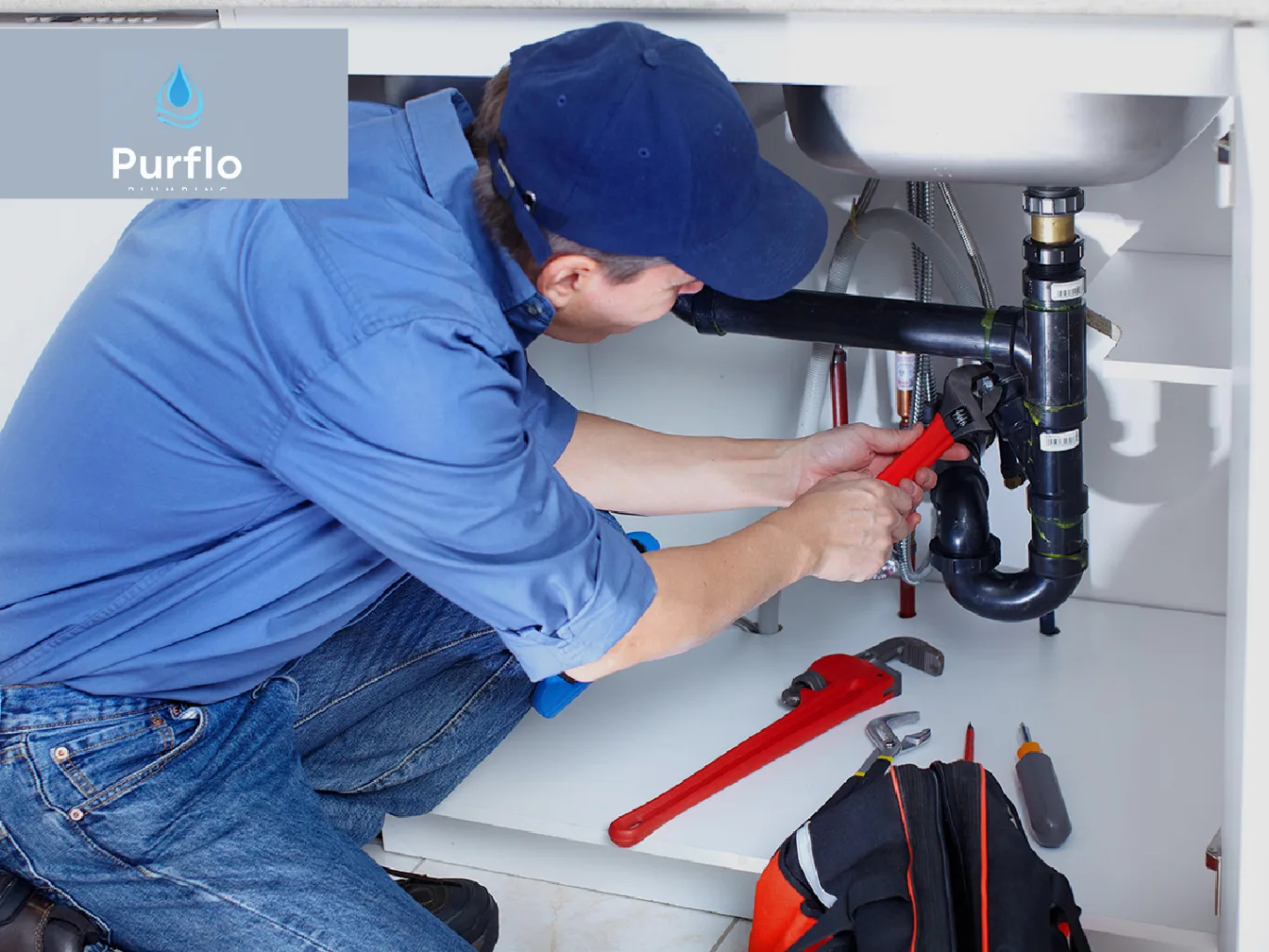 A plumber working on a residential residential plumbing job — mid-task, hands, tools and the work itself in sharp focus, the rest of the space softly out of focus